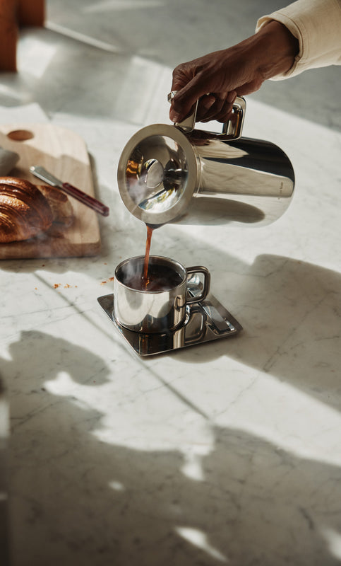 Person pouring coffee from a silver french press into a cup on a marble surface.