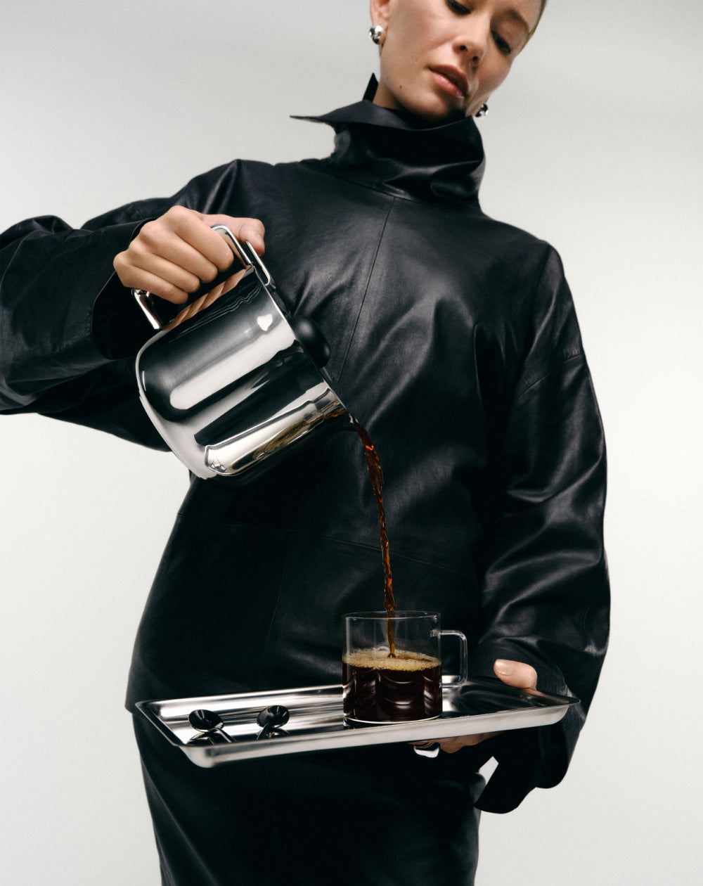 Person in a black outfit pouring coffee from the aarke thermal jug into a glass mug on a white background