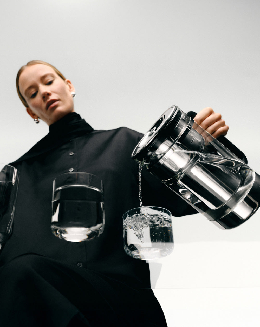 Person holding a the Aarke Water purifier filter pitcher with water being poured into nesting glasses on a white background