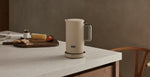 AARKE Kettle Sand electric kettle on a kitchen counter with stainless steel handle, styled in a warm Scandinavian kitchen setting.