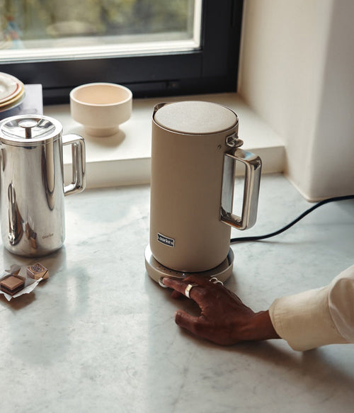 AARKE Kettle Sand electric kettle on kitchen counter by a window, featuring stainless steel handle and Scandinavian design. Perfectly paired with the Aarke Coffee French Press.