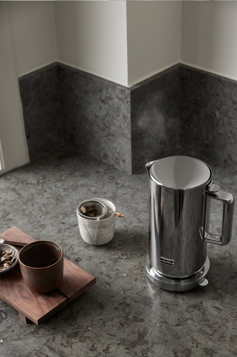 The Aarke Kettle in stainless steel emitting steam on a dark stone kitchen counter next to cups of tea.
