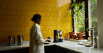 Person standing in a kitchen with yellow tiled walls and various aarke kitchen appliances on the counter.