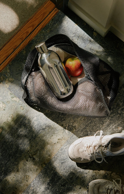 Mesh bag with a to-go bottle in stainless steel and apple on a stone floor next to a white shoe.