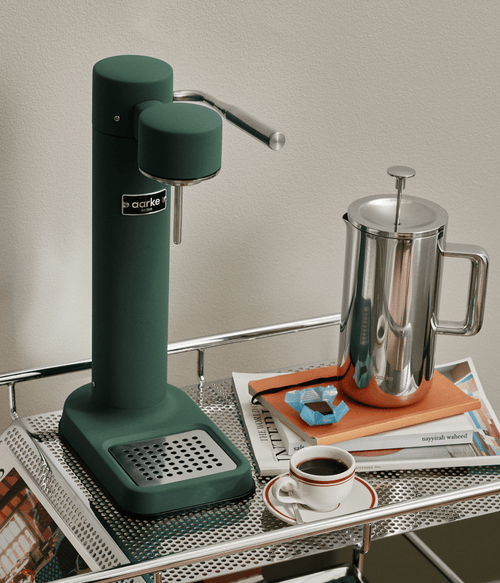 Aarke Carbonator 3 in Heritage Green next to a the Aarke stainless steel coffee press, creating sparkling water in a modern kitchen setting.