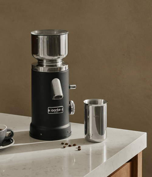 Aarke Coffee Grinder in Matte Black with stainless steel grounds container and coffee beans on a kitchen countertop.