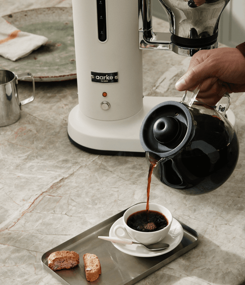Aarke matte white automatic pour-over coffee maker on marble kitchen countertop as freshly brewed coffee is poured from glass carafe into a cup.