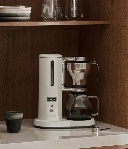 Aarke Coffee Maker in Matte White brewing filter coffee on a marble kitchen countertop with a glass carafe, stainless steel filter basket, and coffee cup in a modern Scandinavian kitchen.