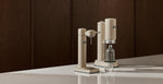 Aarke sparkling water makers in beige on a kitchen countertop with a water bottle and glass, showcasing minimalist Scandinavian design.