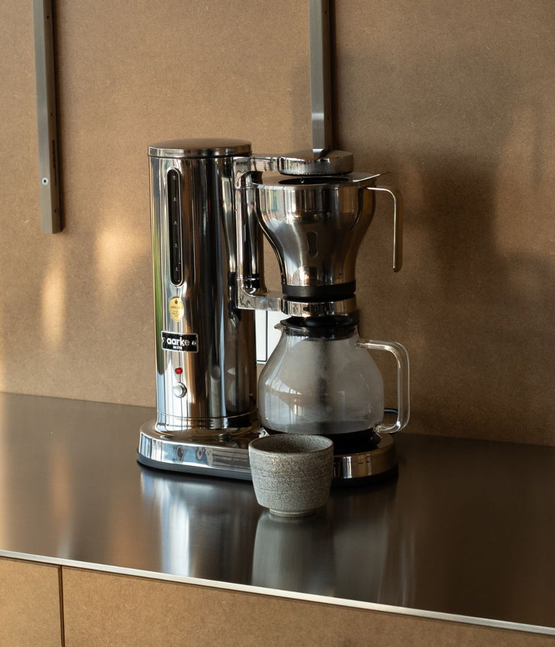 A polished stainless steel Aarke coffee maker with a glass carafe and a ceramic cup beside it, placed on a sleek metal countertop against a wooden wall.