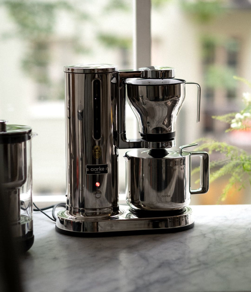 Aarke Coffee Maker brewing directly into the stainless steel Thermal Jug on a marble countertop by a window.